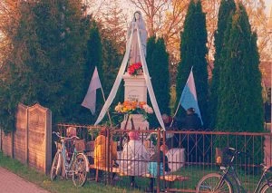 Przydrożna kapliczka z&nbsp;figurą Matki Bożej, kwiatami i&nbsp;flagami.